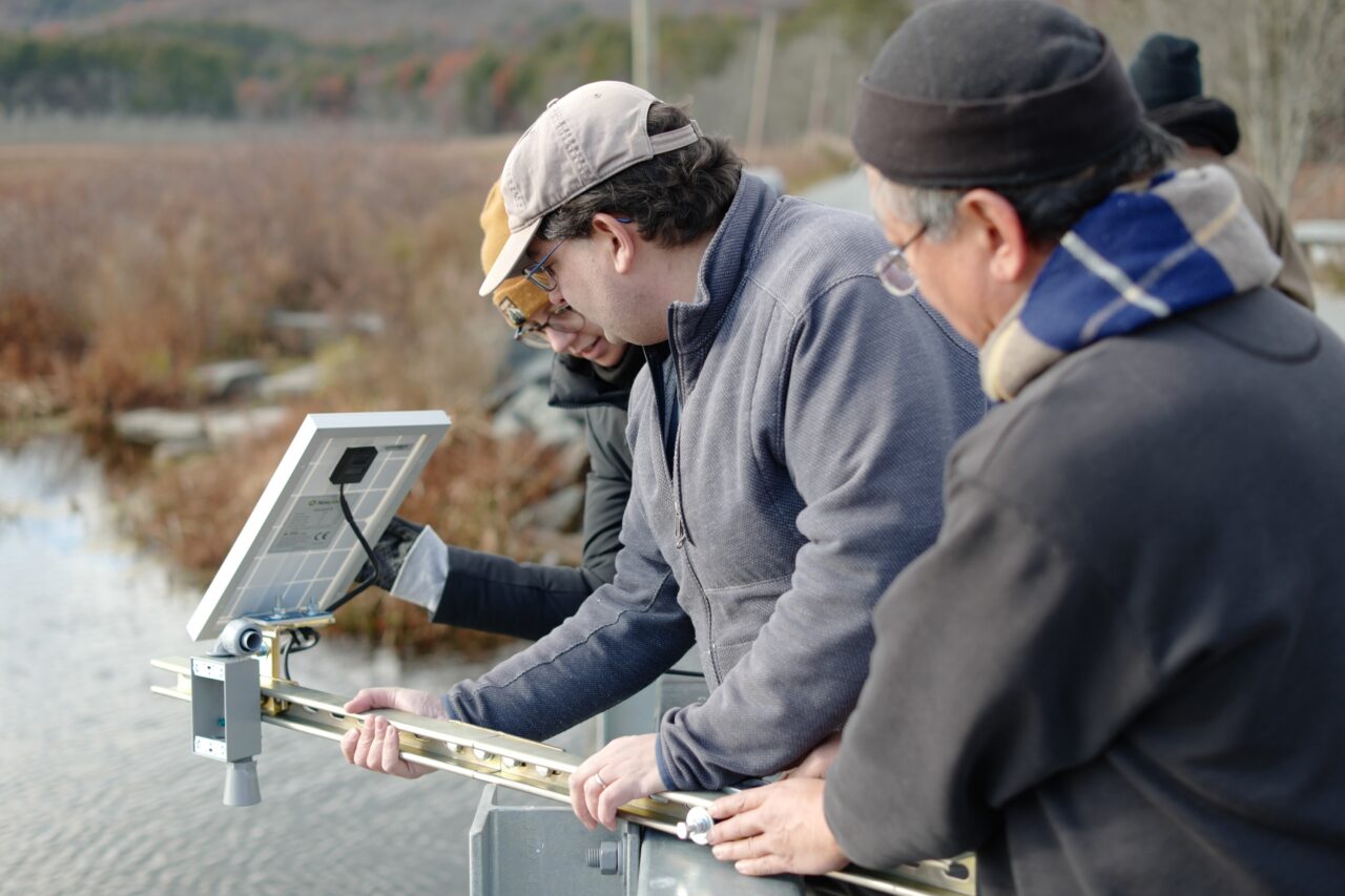 Three people hold the FieldKit sensor mount over the edge of a bridge. The solar panel and ultrasonic rangefinder can be seen.